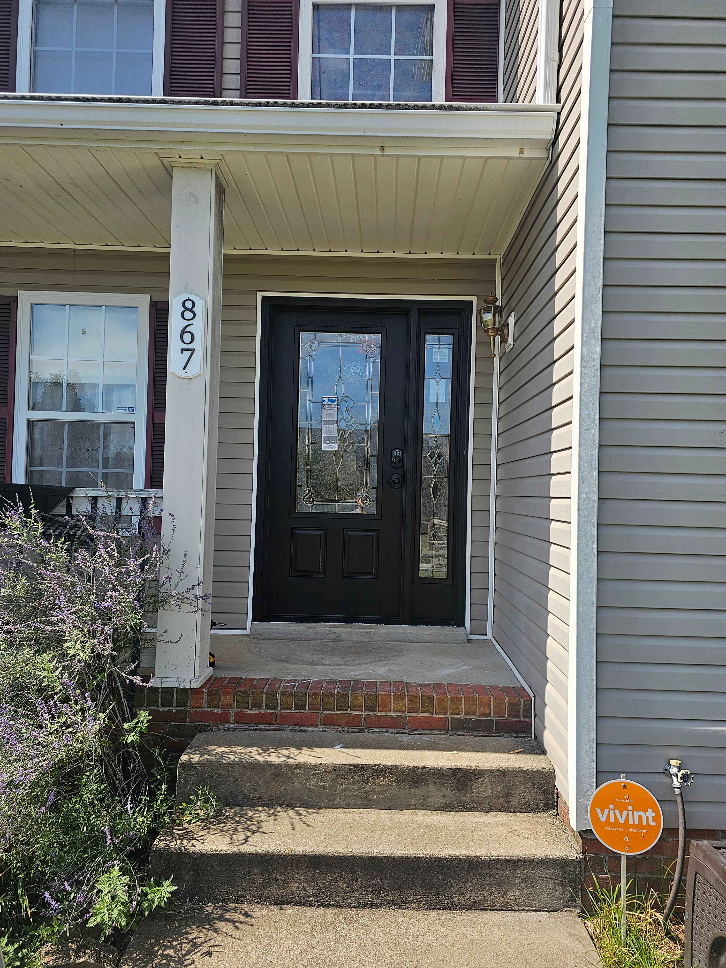Black front door with decorative glass, house number 867, concrete steps, and purple flowering plants.
