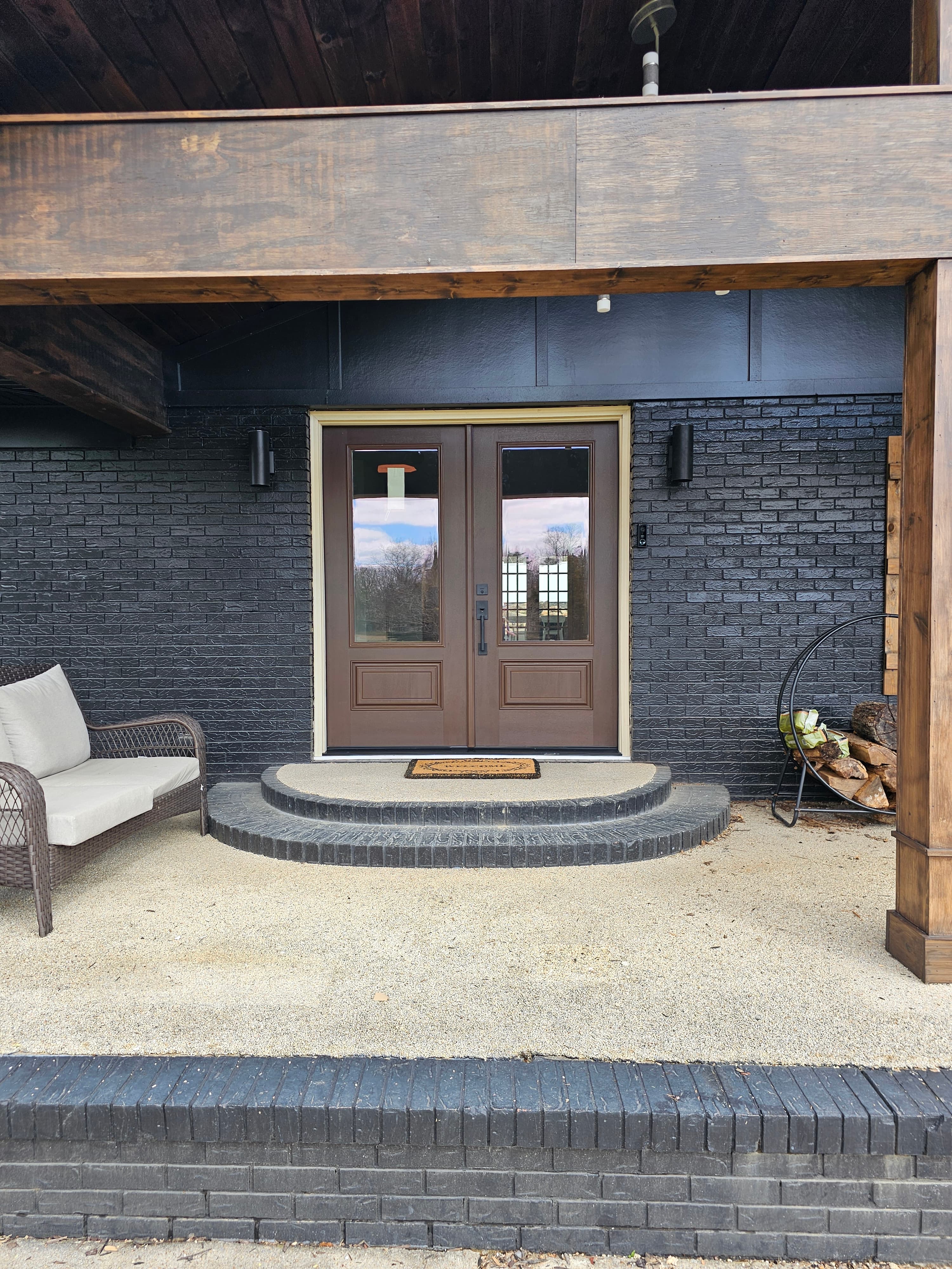Brown double doors on black brick wall with curved stone steps and a wicker chair.